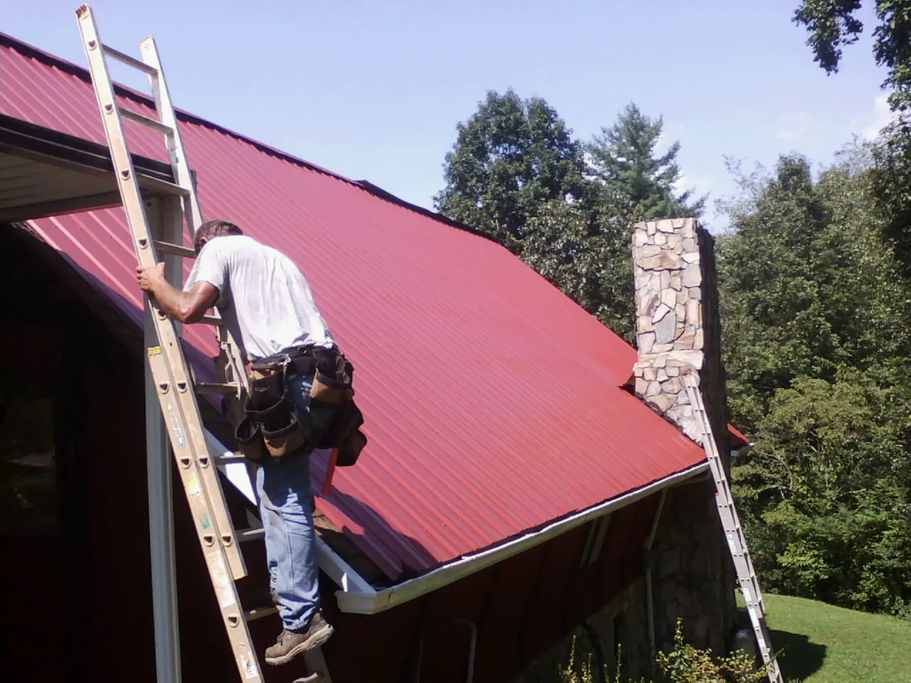 Roofer on a ladder beside a red metal roof and stone chimney — Soffit & Fascia Repair by Roofing Brothers in Elko New Market